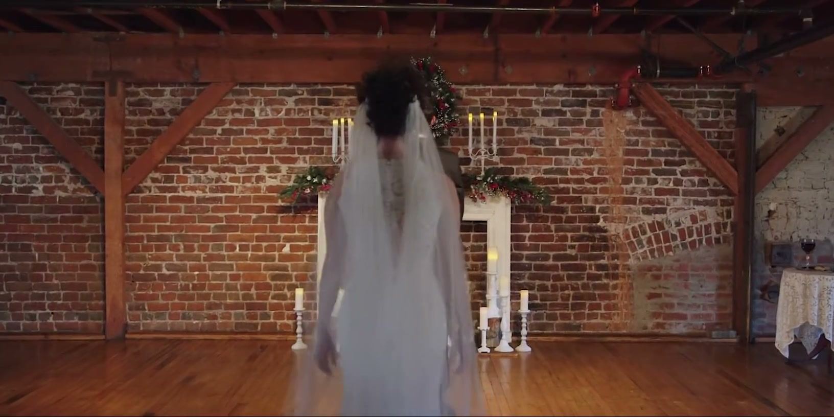A bride seen from behind, walking toward a white mantel set with candelabras and a wreath on the brick wall.