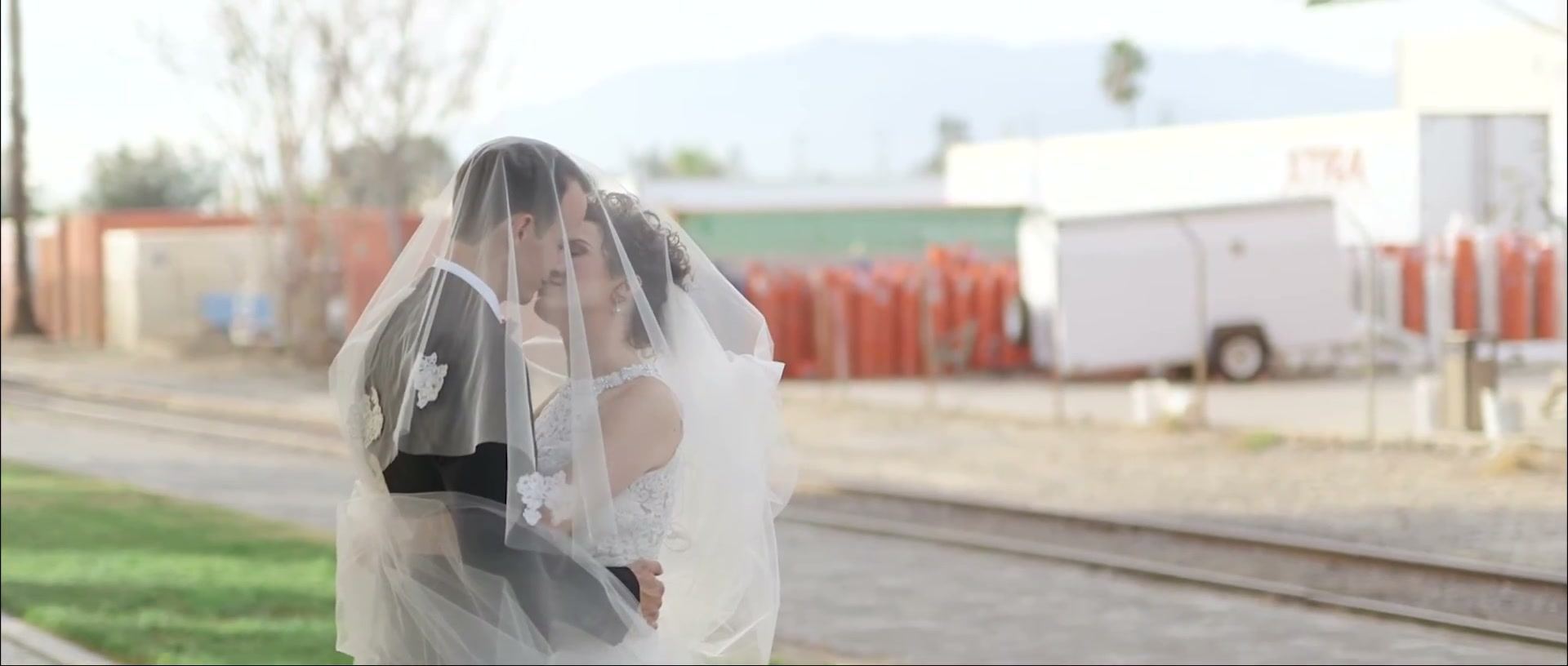 Hayley and Brandon under her veil beside railway tracks, a white trailer and orange barriers behind them.
