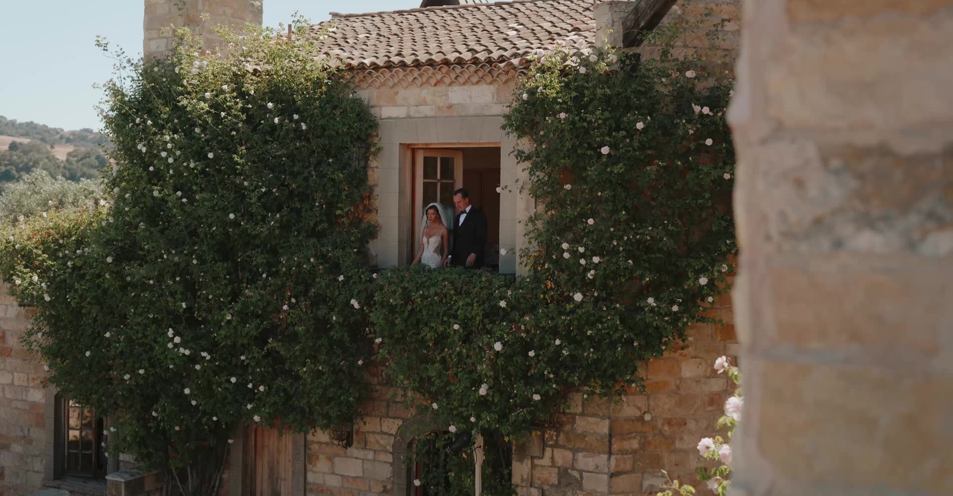 A couple on a villa balcony framed by climbing roses, the opening shot of a Rayne wedding film