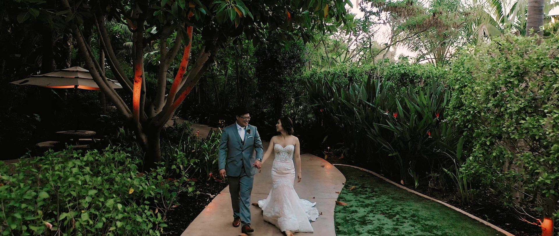 Hugo and Helen walking the curved garden path hand in hand, her train trailing behind her, uplit trees on either side.