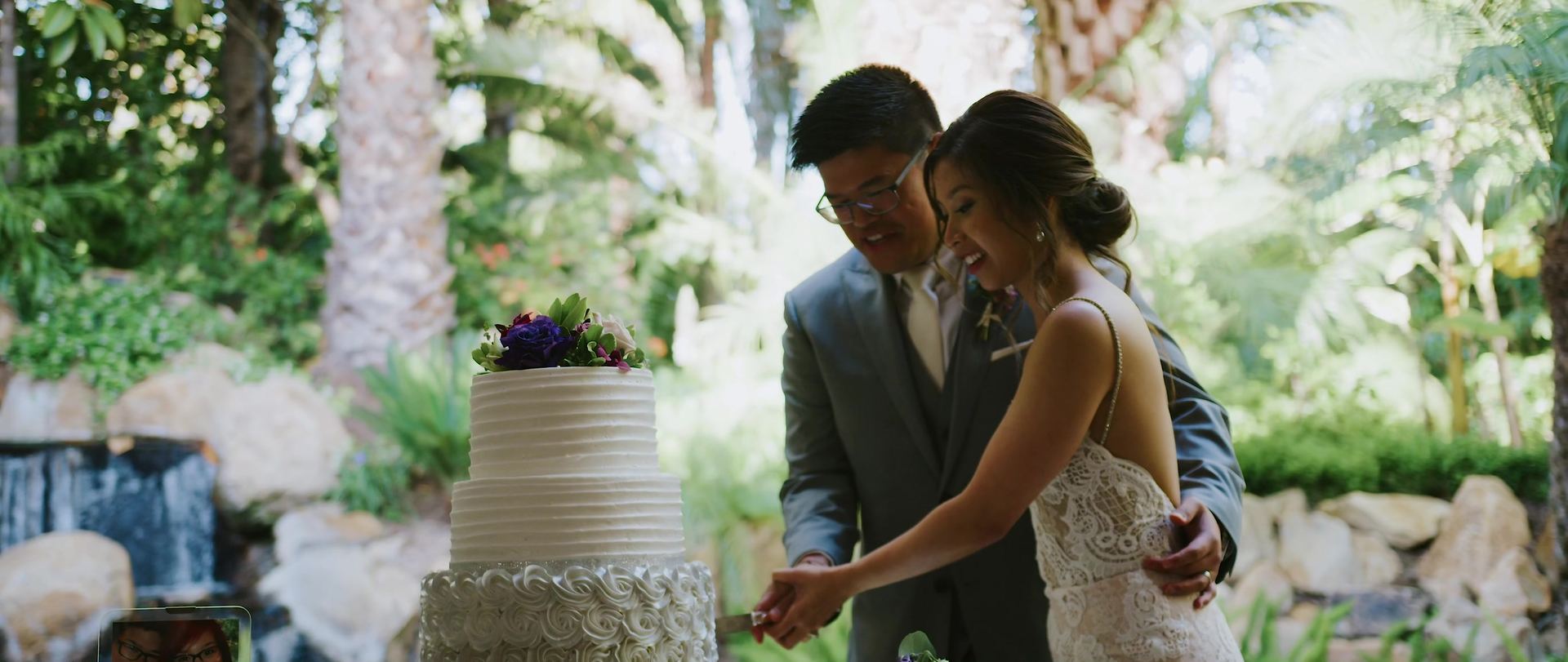 Cutting the cake beside the estate's waterfall, the rock face and greenery rising behind them.