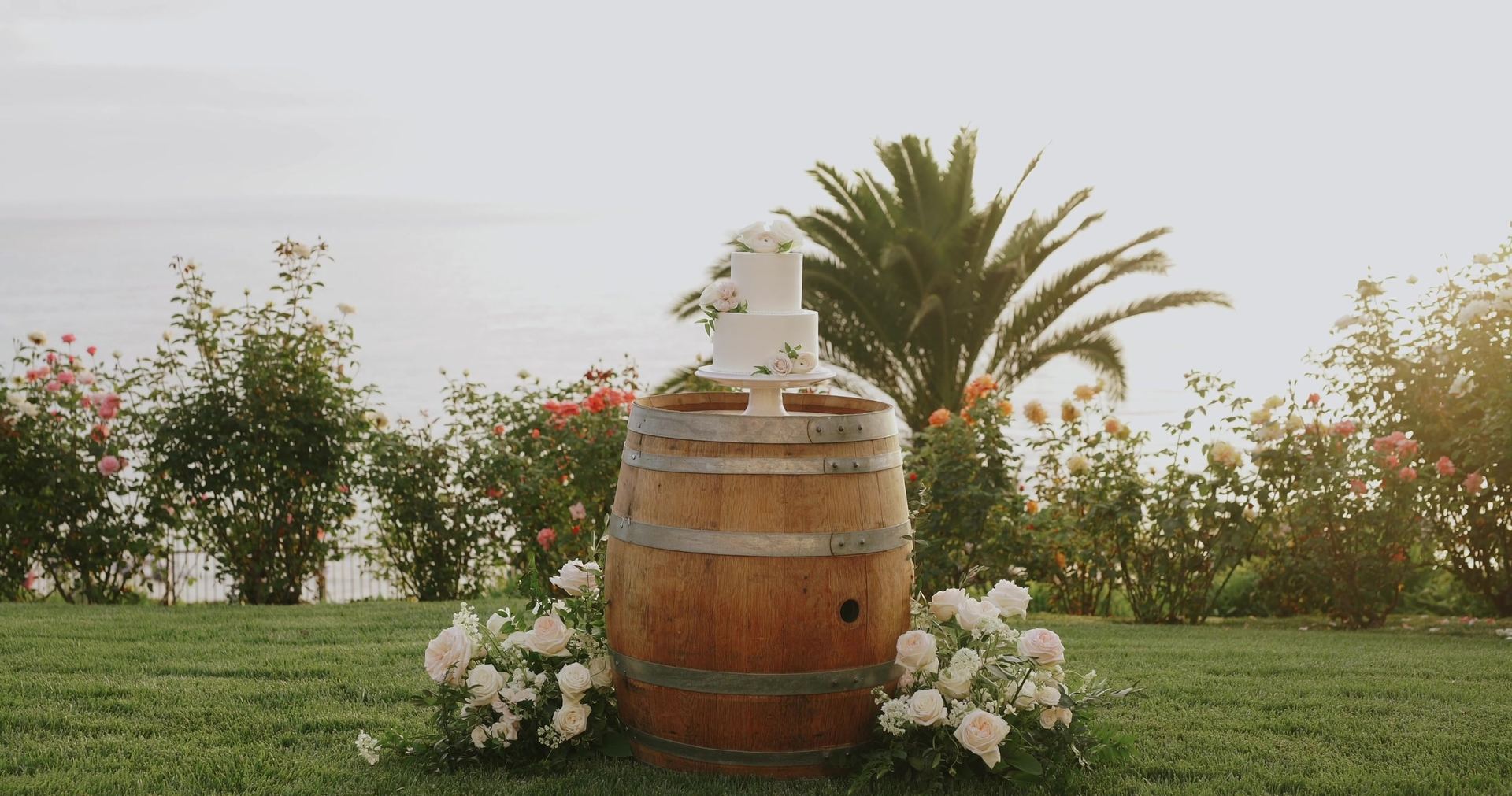The cake set on a wine barrel at the edge of the lawn, white blooms banked around its base.