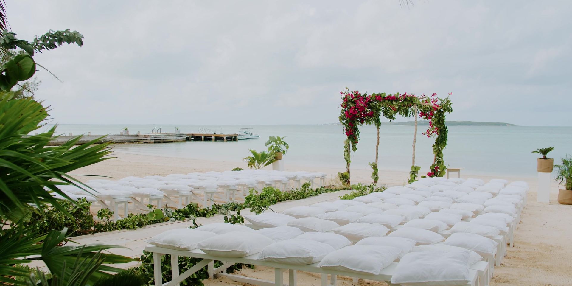 The ceremony set on the sand, a four-post arch draped in bougainvillea and rows of white benches with cushions facing pale water under an overcast sky.