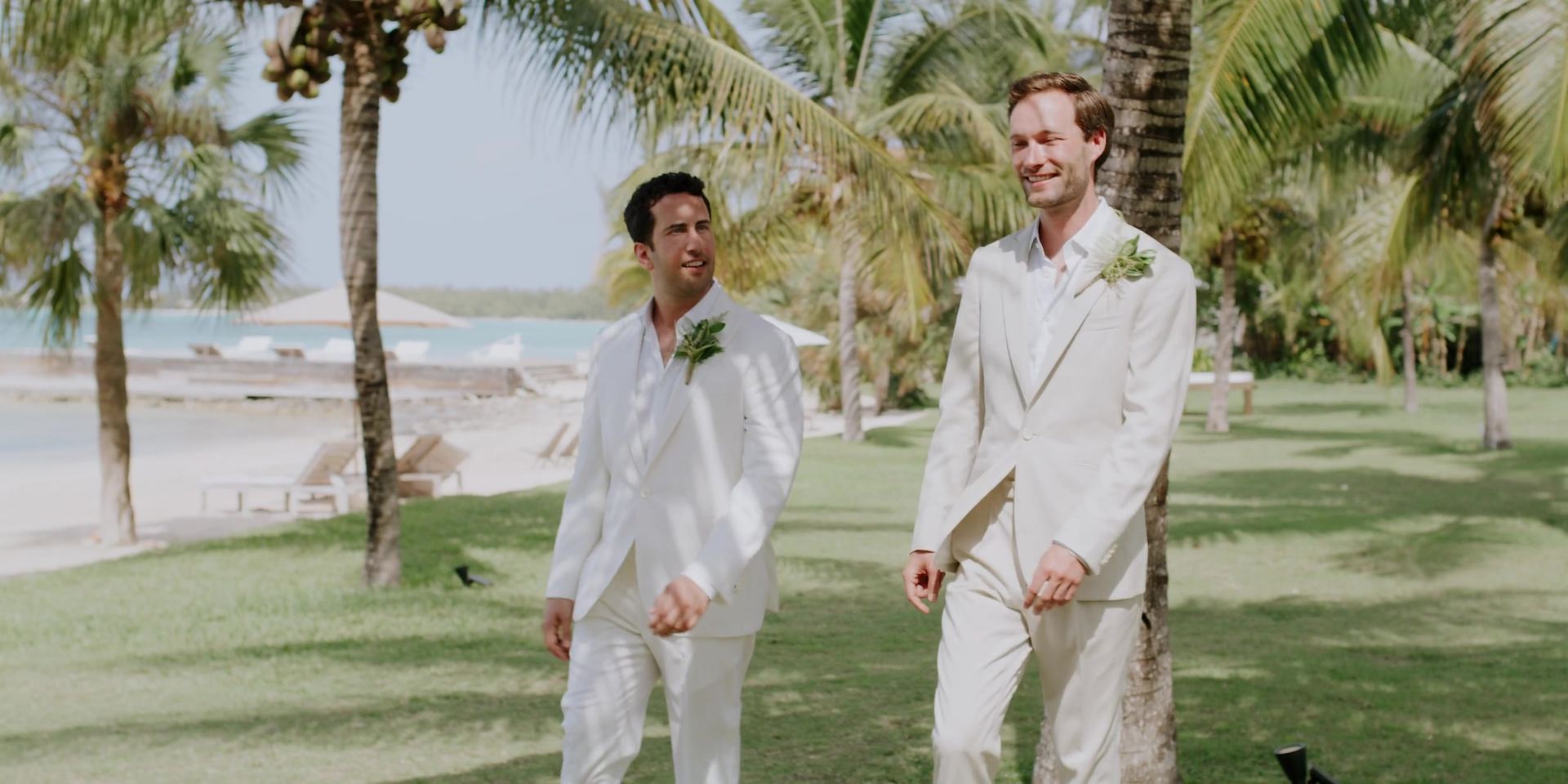 Izak and Slater walking across the lawn under the palms in cream suits, the water and loungers behind them.