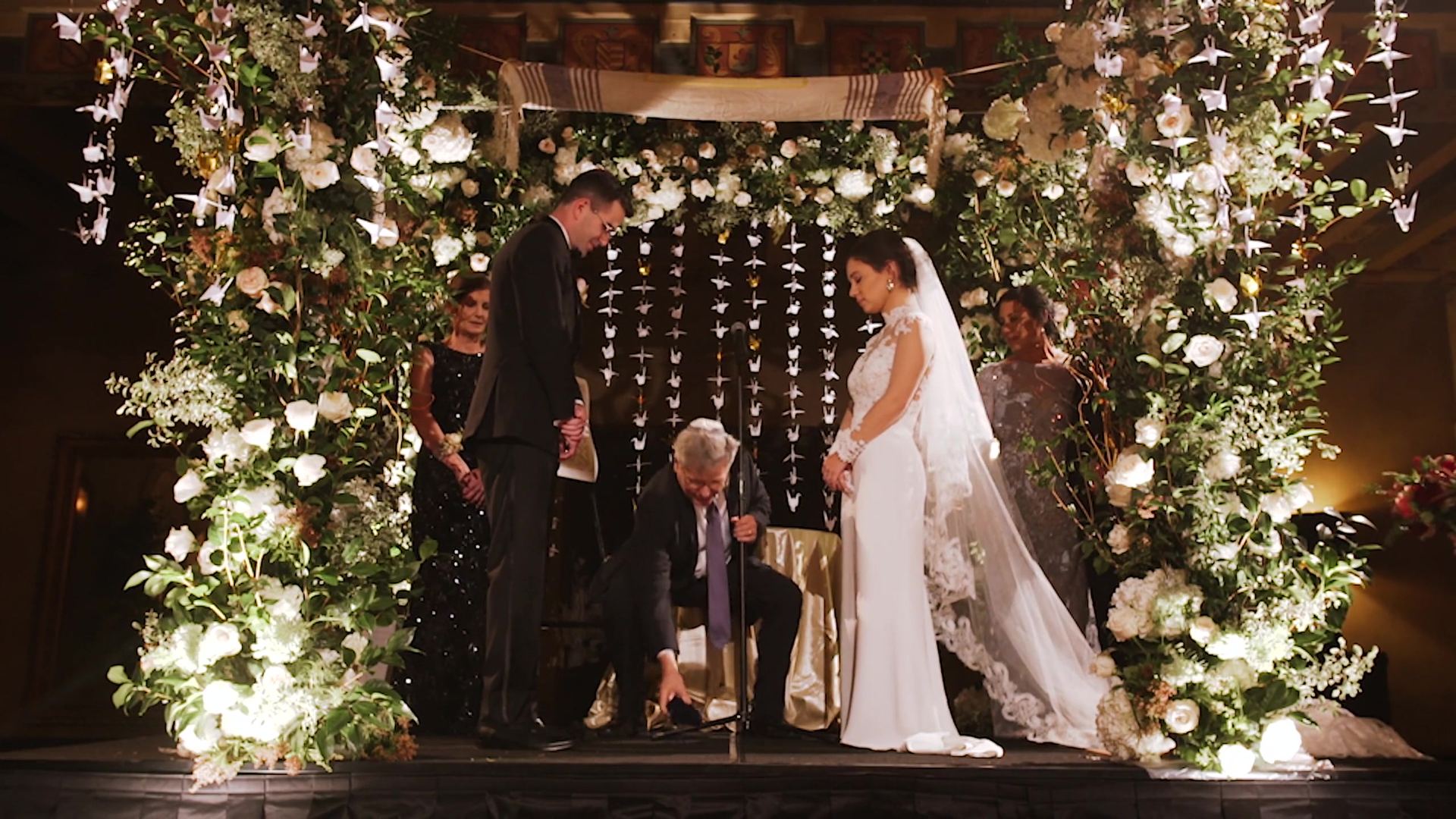 Jordon and Annie under the chuppah, a floral arch of white blooms and greenery with strands of paper cranes hanging behind them, a man crouched between them.