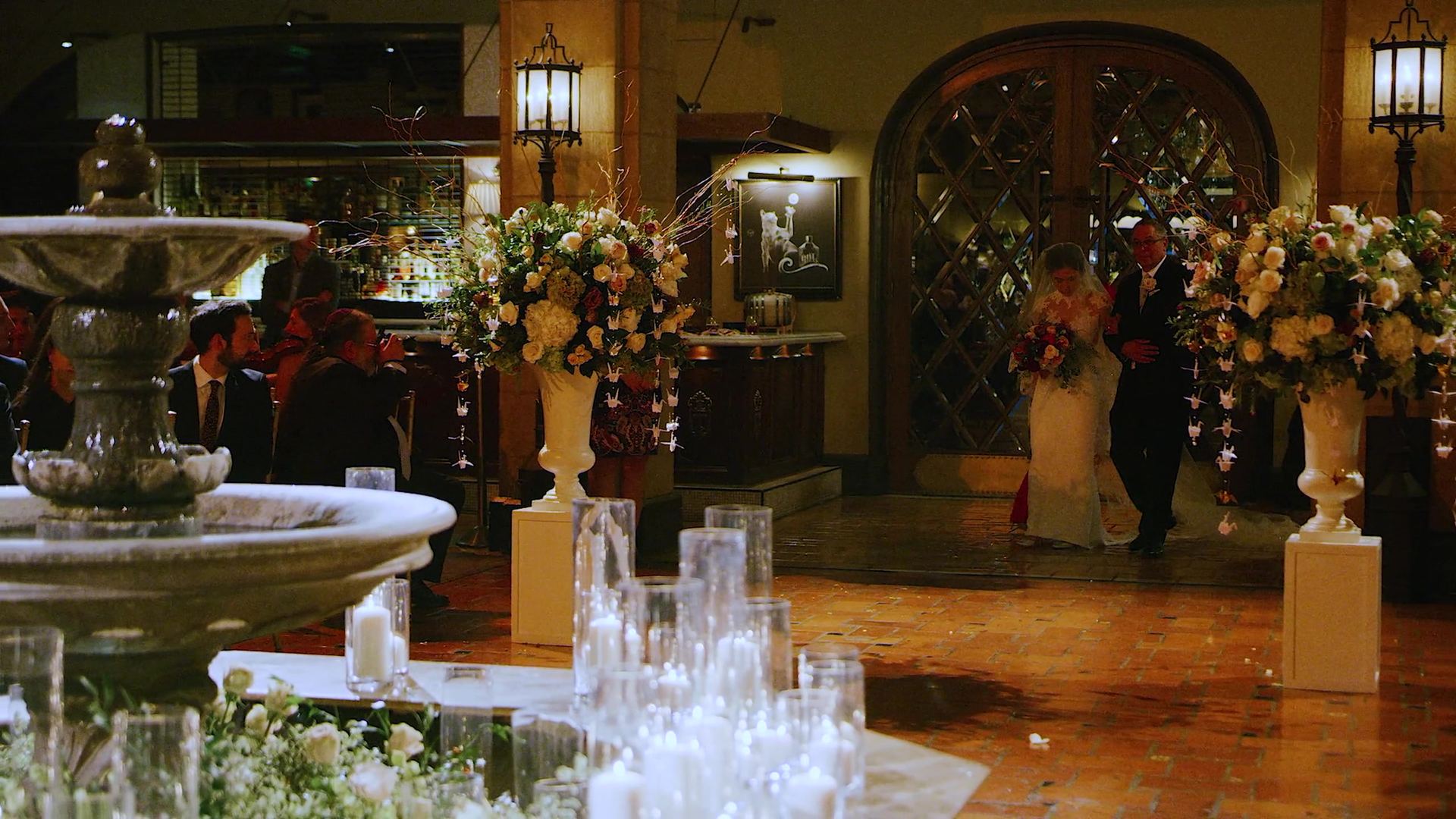 Annie coming through the arched lattice doorway on a man's arm, past tall urns of cream and blush florals, the tiered stone fountain and banks of candles in the foreground.