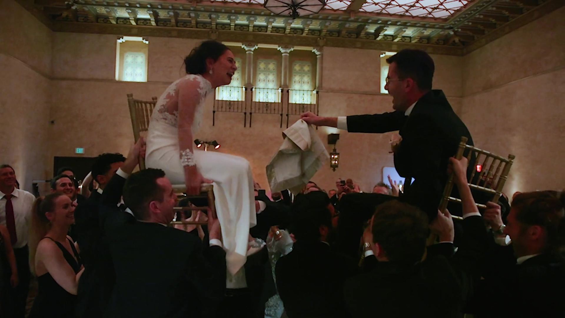 Annie lifted in a chair during the hora, guests crowding in around her, both of them lifted in chairs with a white napkin stretched between them.
