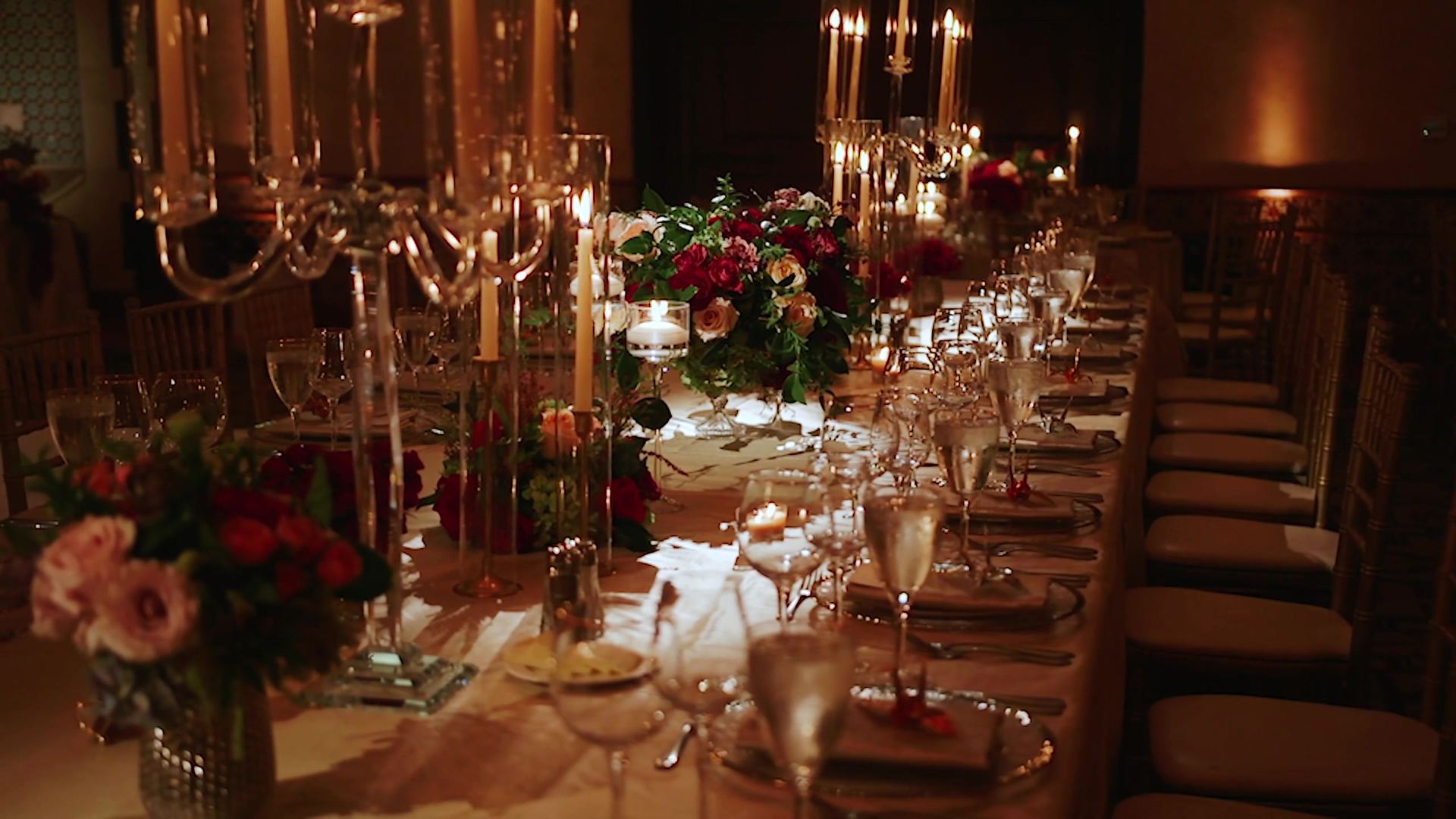 The long reception table, tall glass candelabra with taper candles and low arrangements of red and blush roses down its length, gold chairs along one side.