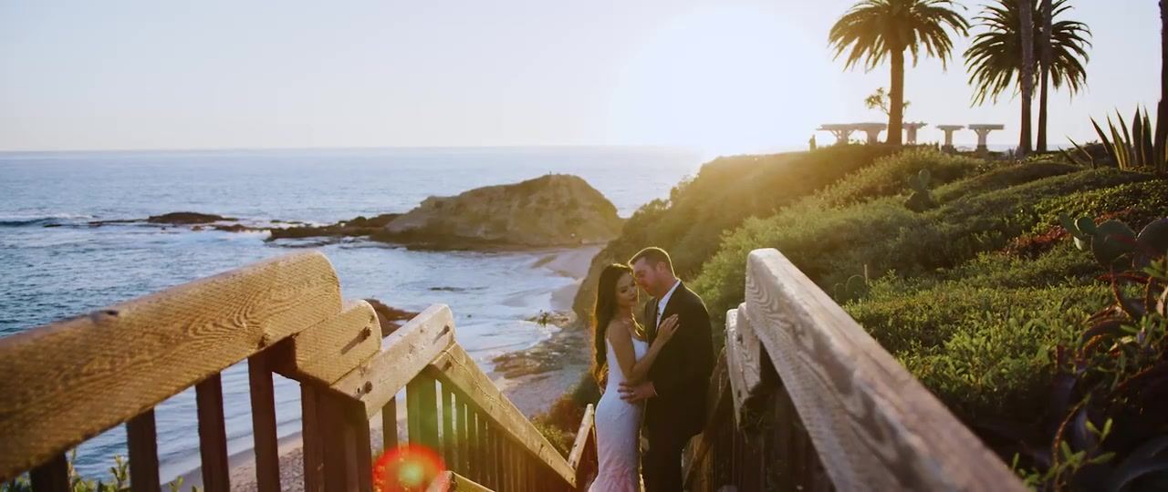 Jan and Brian on the wooden stairway above the cove, palms above them and the sun low over the water.