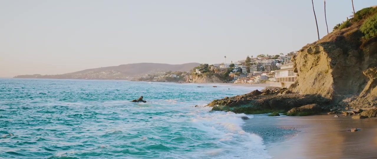 Turquoise water breaking on the rocks below a cliff, houses stacked along the hillside beyond.