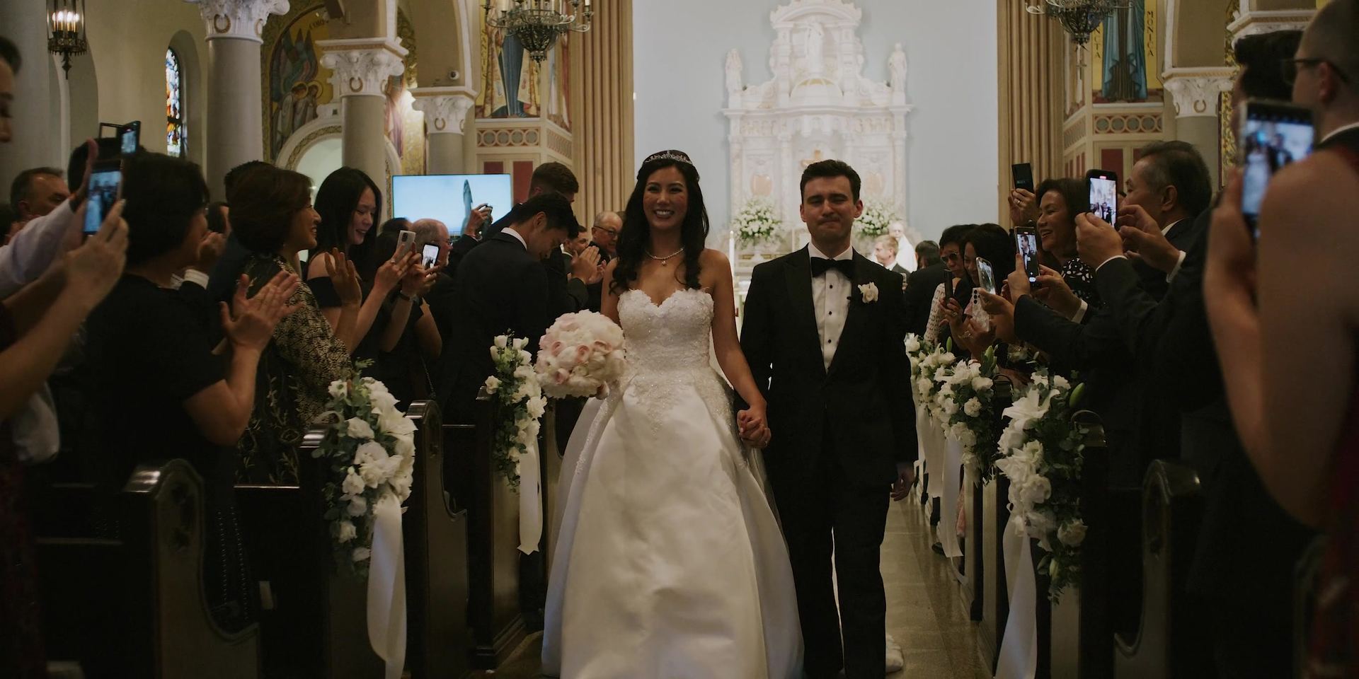 Joe and Jessica coming back down the church aisle, guests leaning out to photograph them.