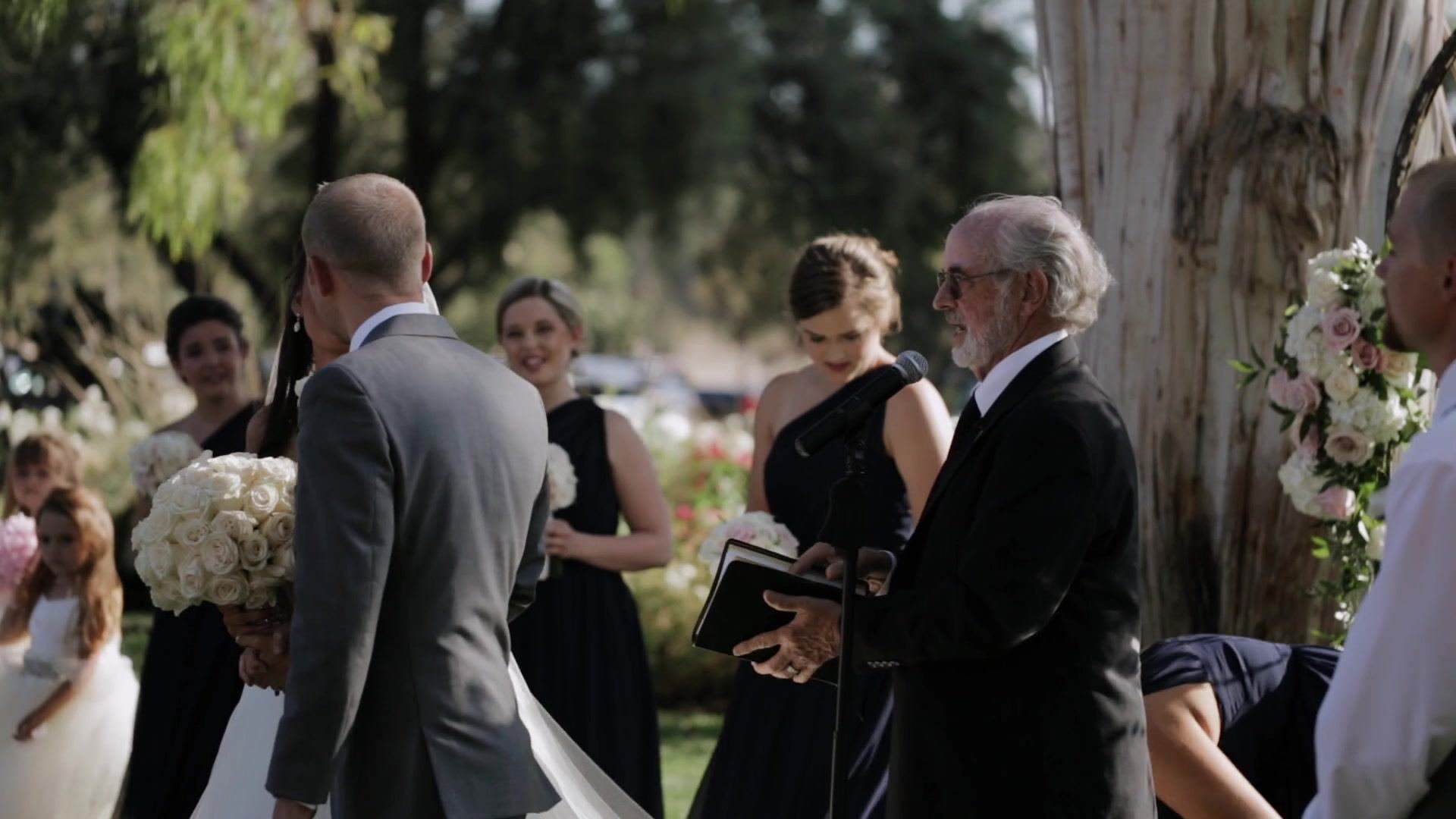 Josh from behind at the altar as the officiant reads from a book, bridesmaids in navy and a flower girl to one side.