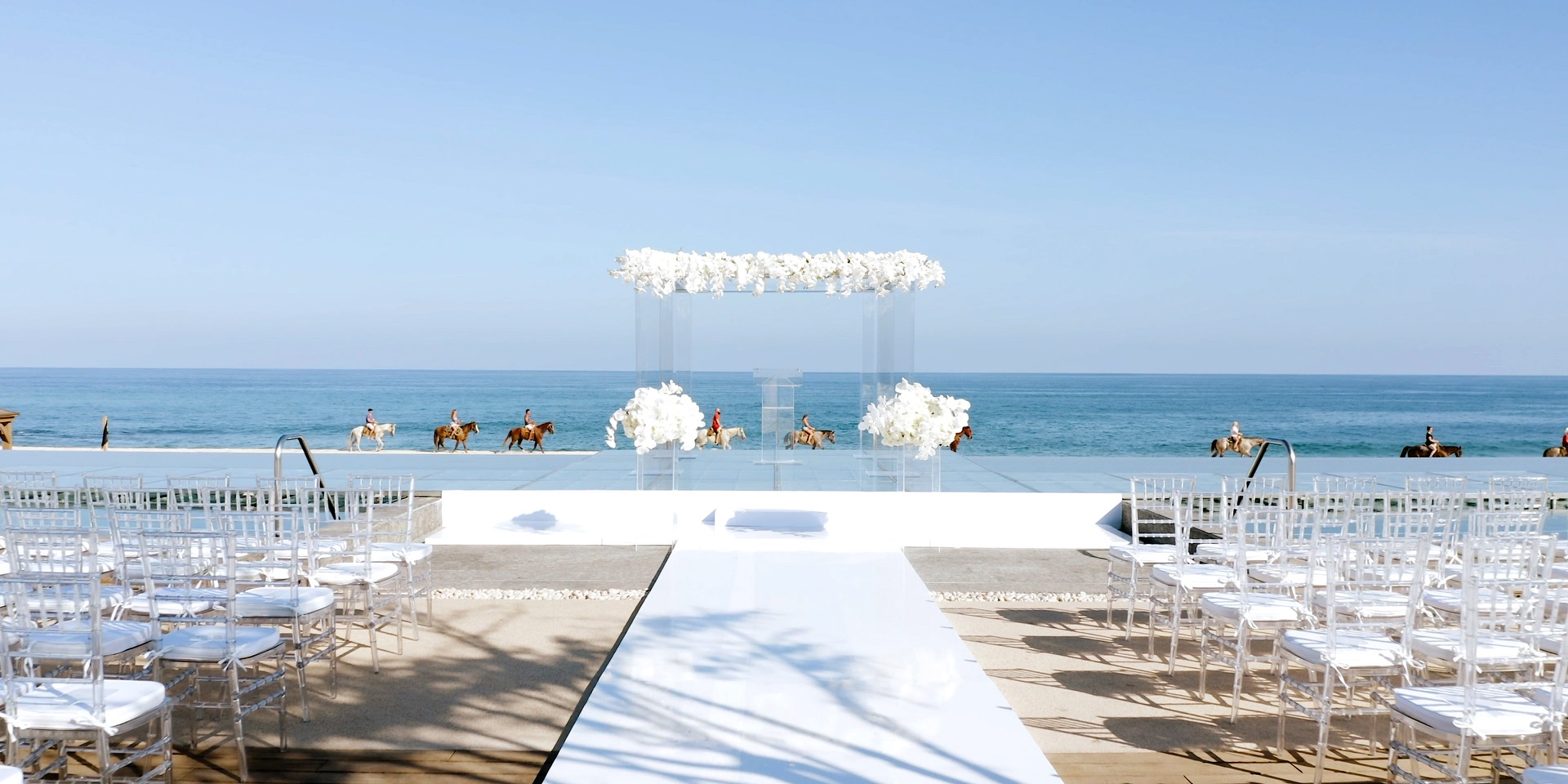Oceanfront wedding ceremony setup in San Jose del Cabo, an acrylic chuppah with white florals facing the Sea of Cortez as horseback riders pass on the beach