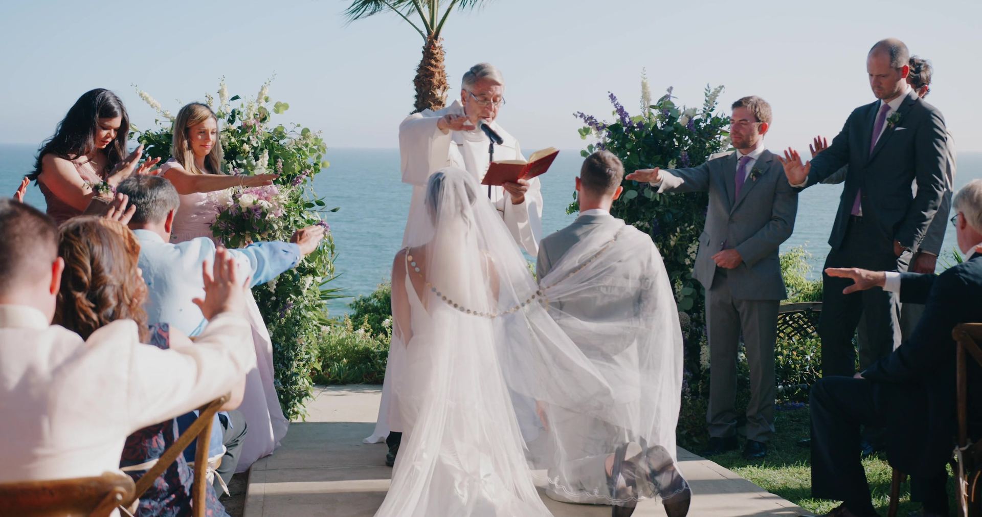 James and Katrina kneeling under a veil and cord as the officiant reads, the wedding party and guests stretching their hands toward them, the ocean behind the altar.