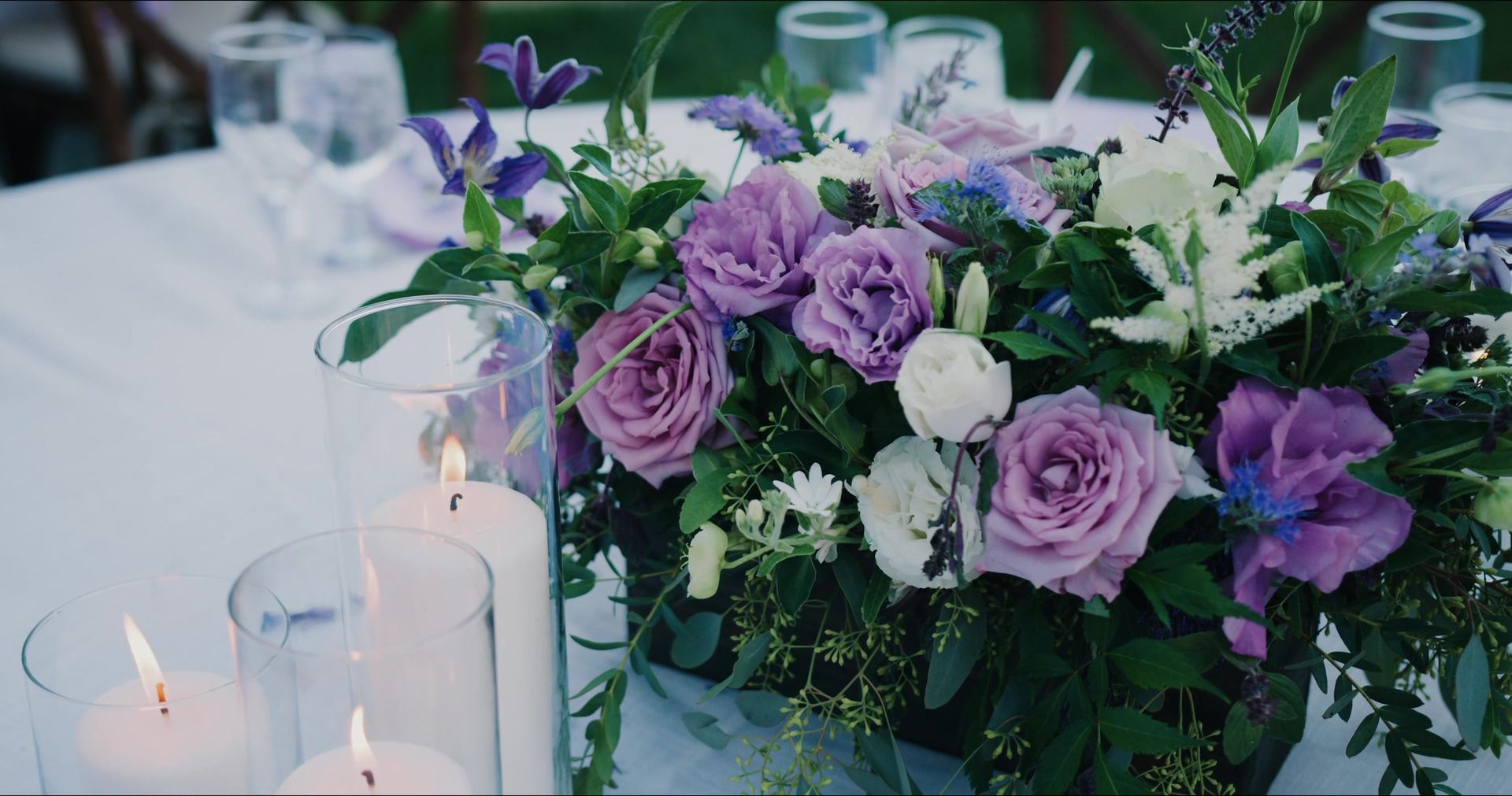 A centrepiece of purple roses and lisianthus beside pillar candles on the table.