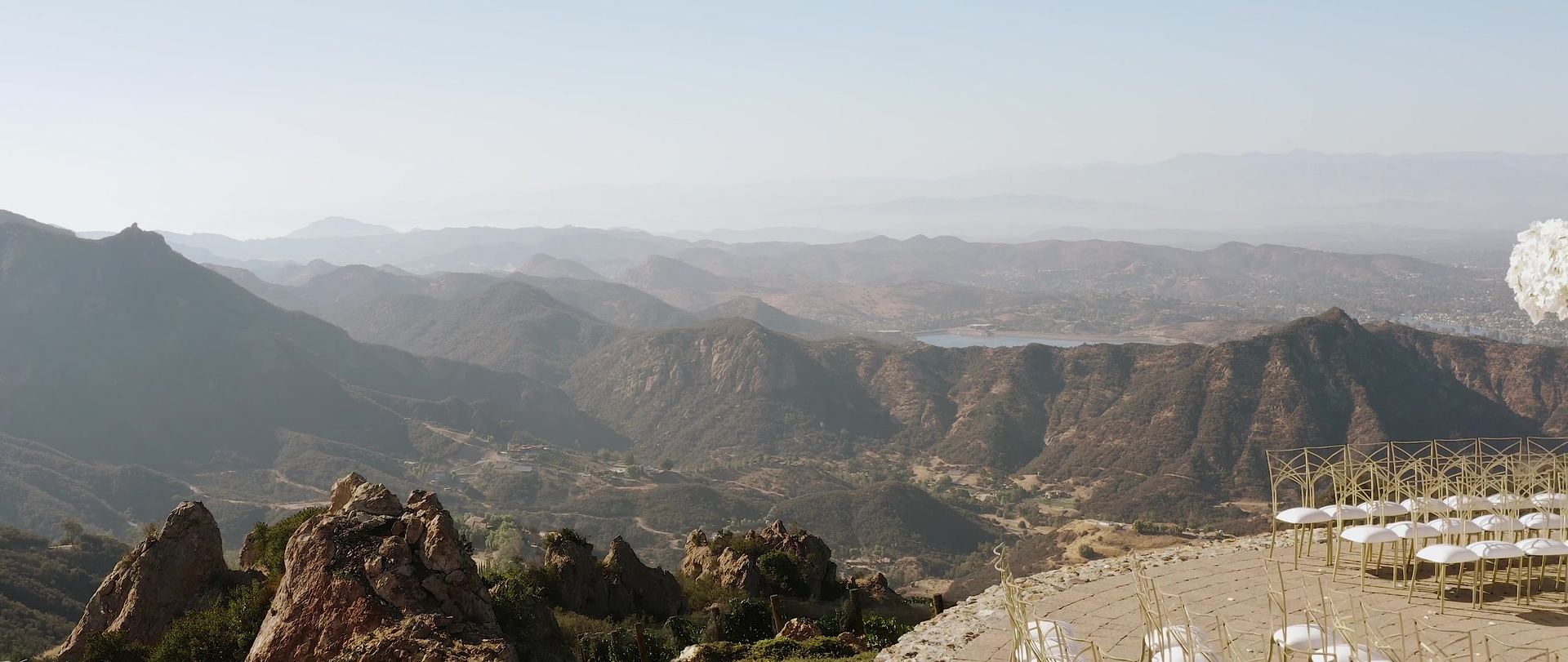 The view from Malibu Rocky Oaks, ridgelines receding into haze with a lake in the middle distance, and rows of gold-framed chairs on the stone ceremony terrace at the right.