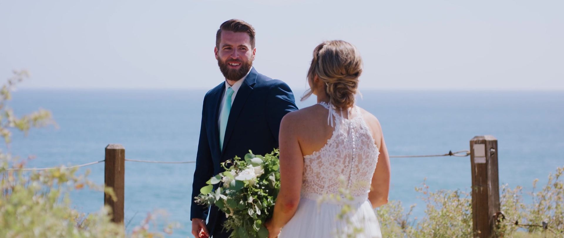 Josh turning to see Shayna for the first time at the rope fence above the water, her bouquet of white blooms and greenery in hand.