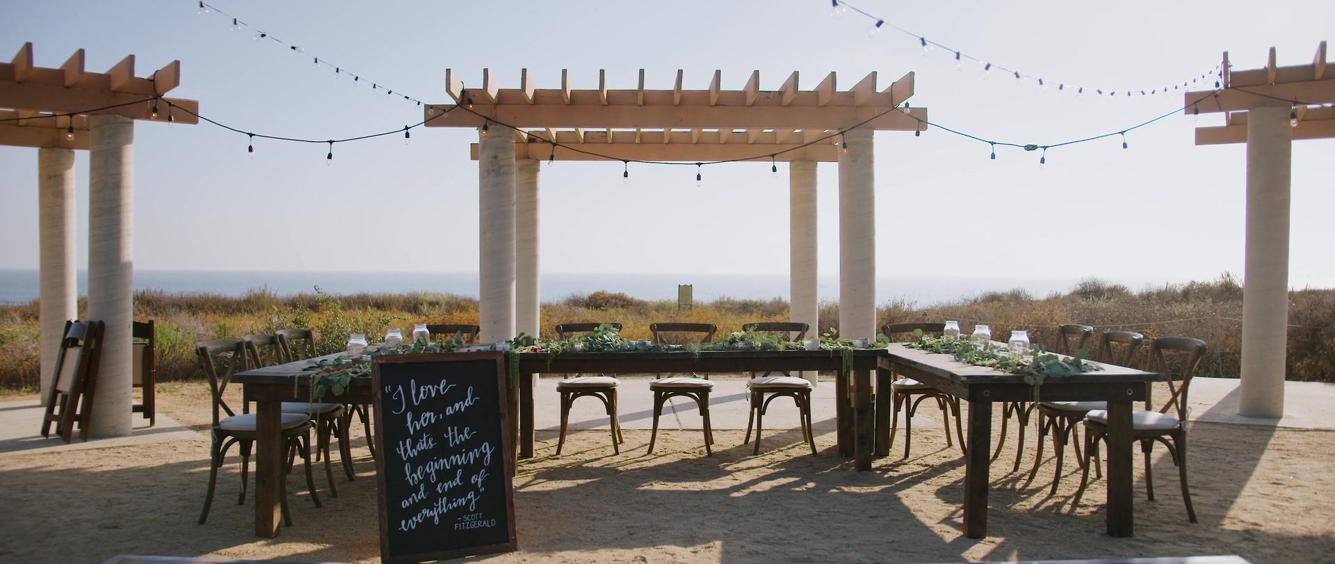Timber pergolas on concrete columns strung with lights, long wooden tables set with greenery beneath them and the bluff falling away to the water behind.