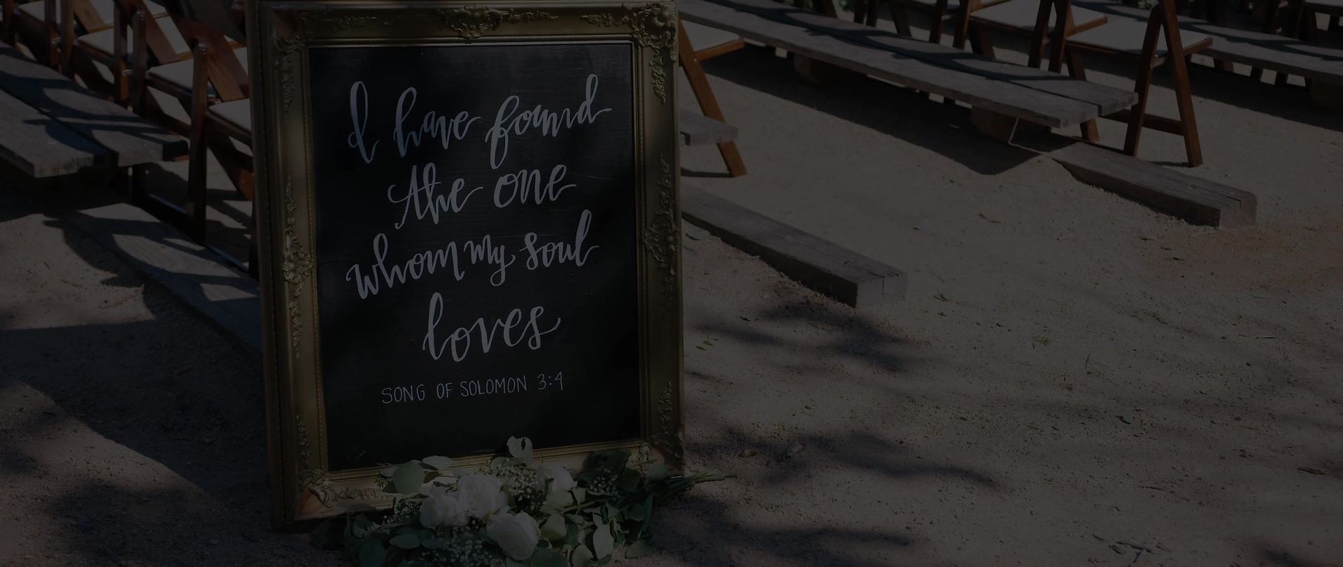 A gold-framed chalkboard on the sand reading I have found the one whom my soul loves, Song of Solomon 3:4, wooden benches set behind it.