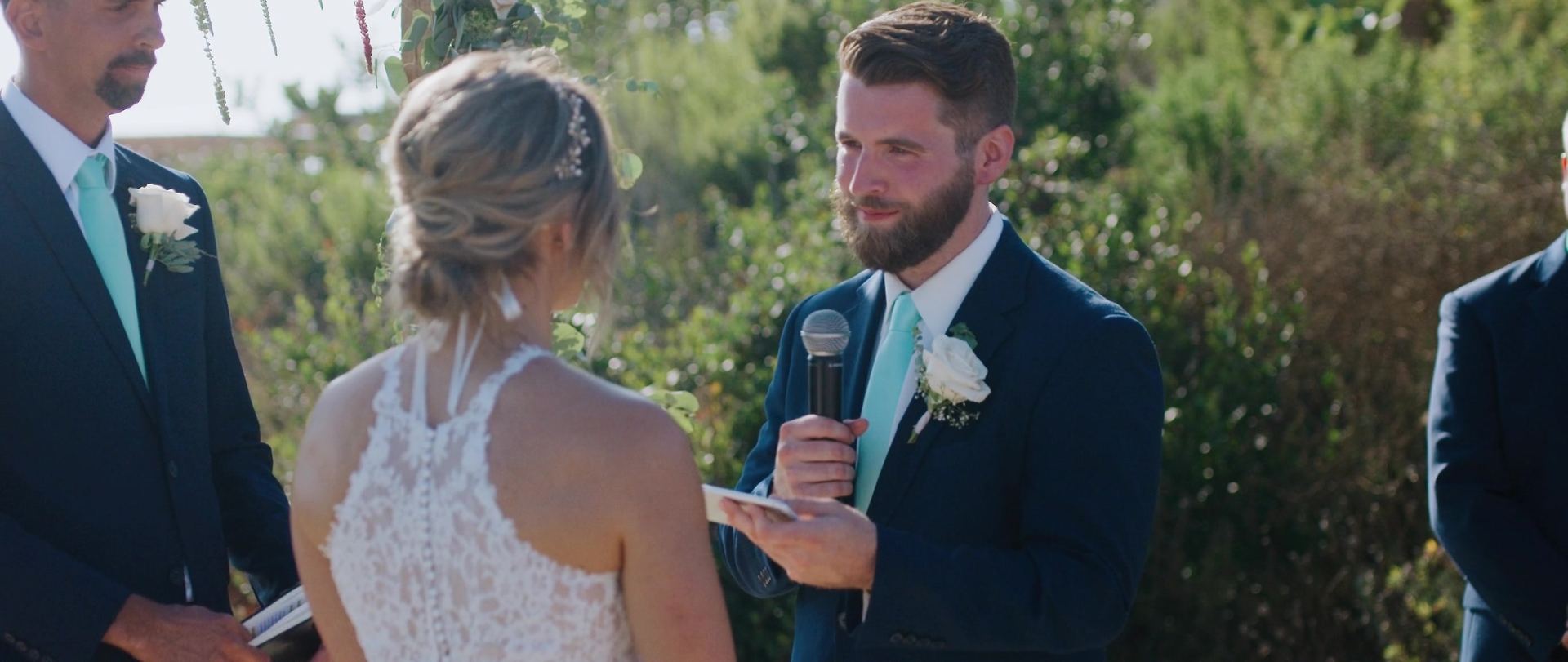 Josh reading his vows into a microphone, Shayna facing him, a groomsman in a mint tie beside them.