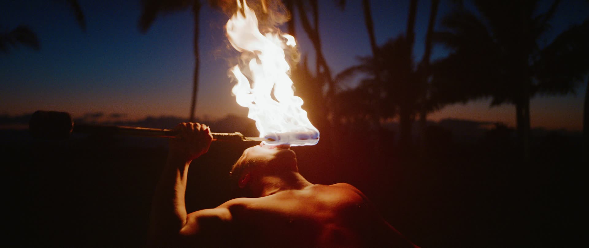 A torch being lit at dusk against silhouetted palms at Paradise Cove, Oahu
