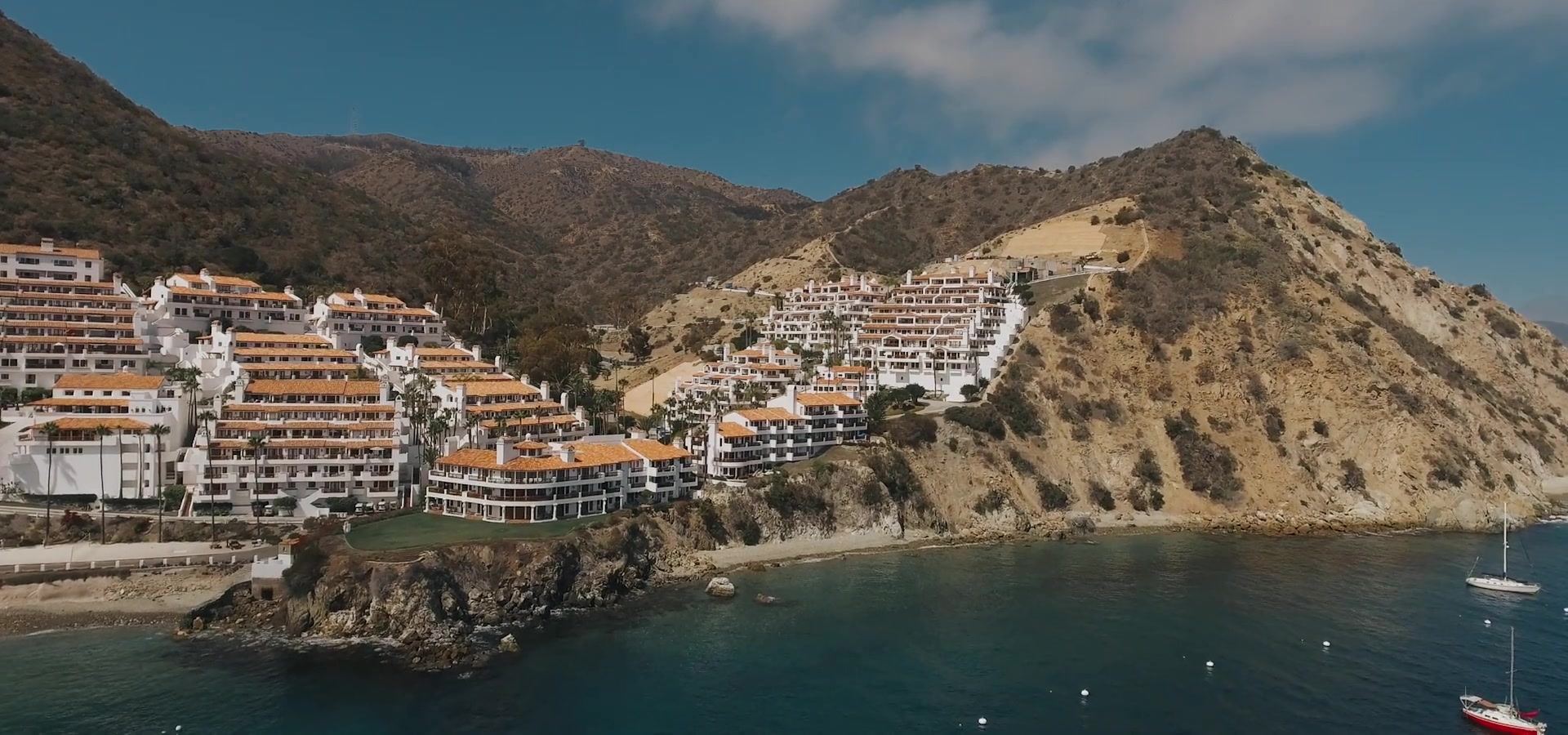 White buildings with terracotta roofs stacked up a dry hillside above a cove, sailboats moored below.