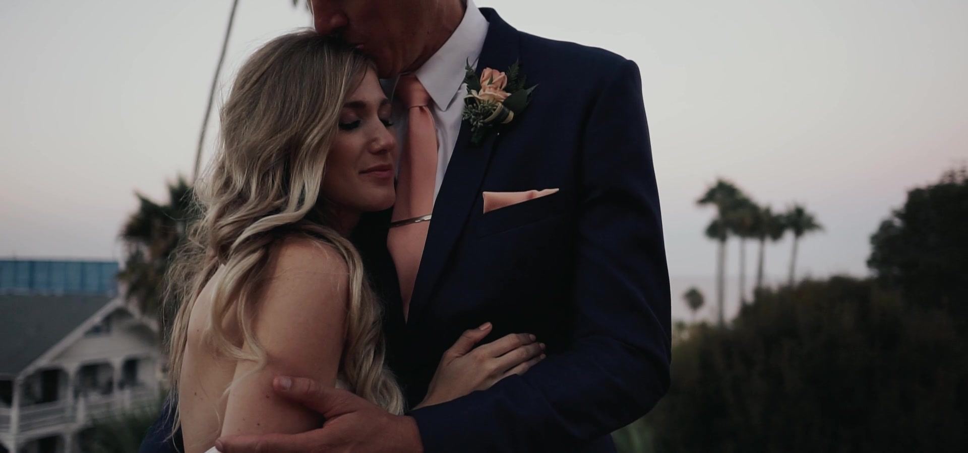Kyle kissing Ava's forehead, palms and a white balconied building behind them under a pale sky.