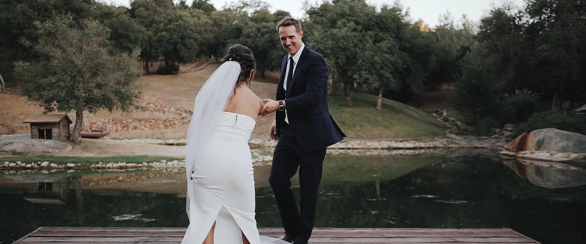 Kevin taking Keshiia's hand as she steps onto the wooden dock at the pond, oak-dotted hills on the far bank.