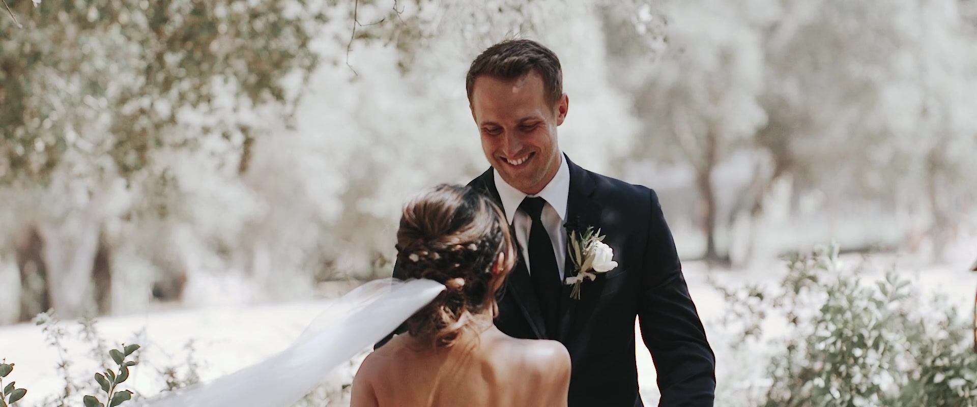 Kevin smiling down at Keshiia as she reaches him, her veil pinned into her hair, trees bright with backlight behind them.