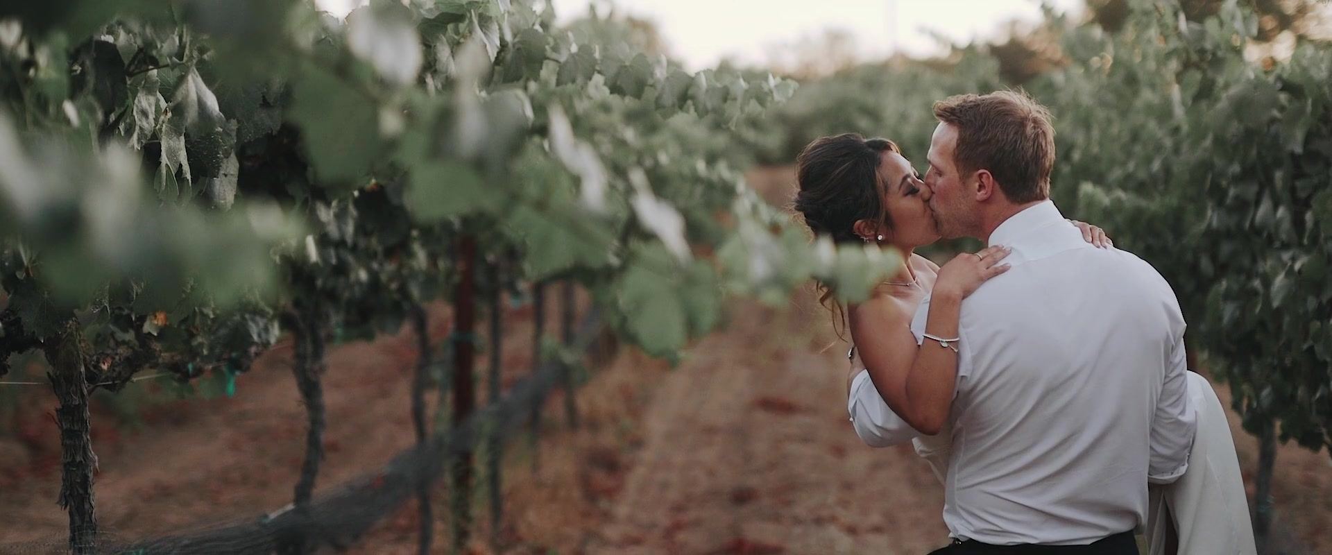 Kevin and Keshiia kissing between the vine rows, his jacket off, her hand on his shoulder.