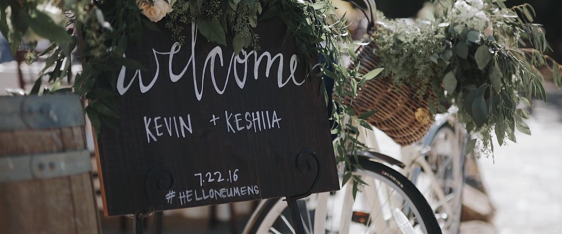 A chalkboard reading welcome Kevin + Keshiia 7.22.16 #helloneumens, propped against a bicycle with a basket of greenery beside a wine barrel.