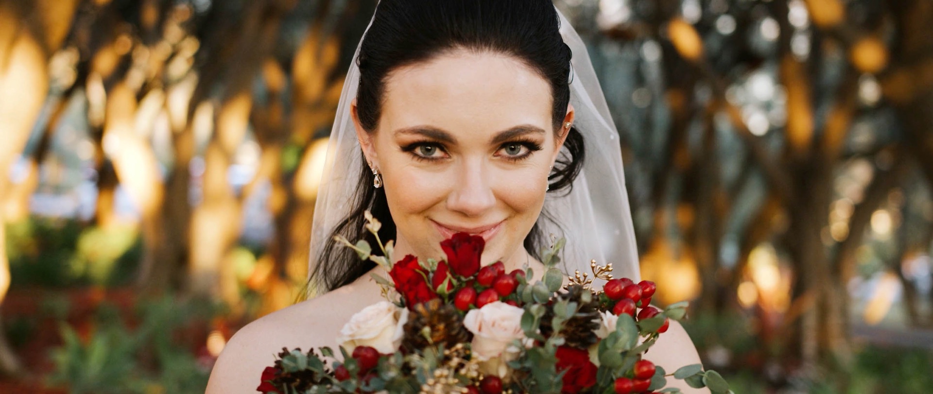 Bride at Padua Hills Theatre holding a red rose bouquet, the olive grove glowing gold behind her
