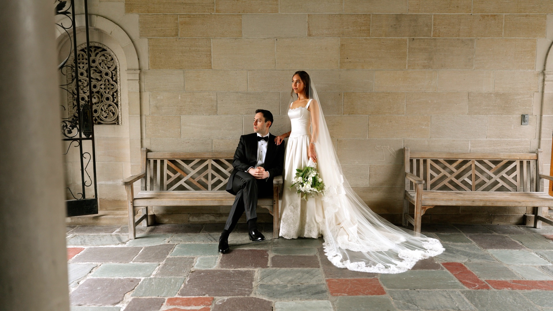 A bride and groom on a stone loggia with a wooden bench and ornate ironwork, on a rainy Los Angeles wedding day