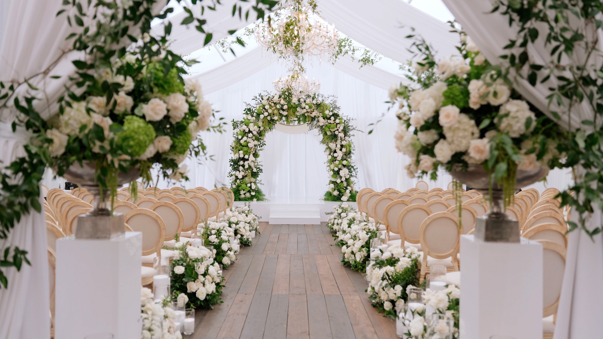 A Maybourne Beverly Hills wedding ceremony under a clear-roofed tent, white drape and a floral chuppah at the end of a wooden aisle