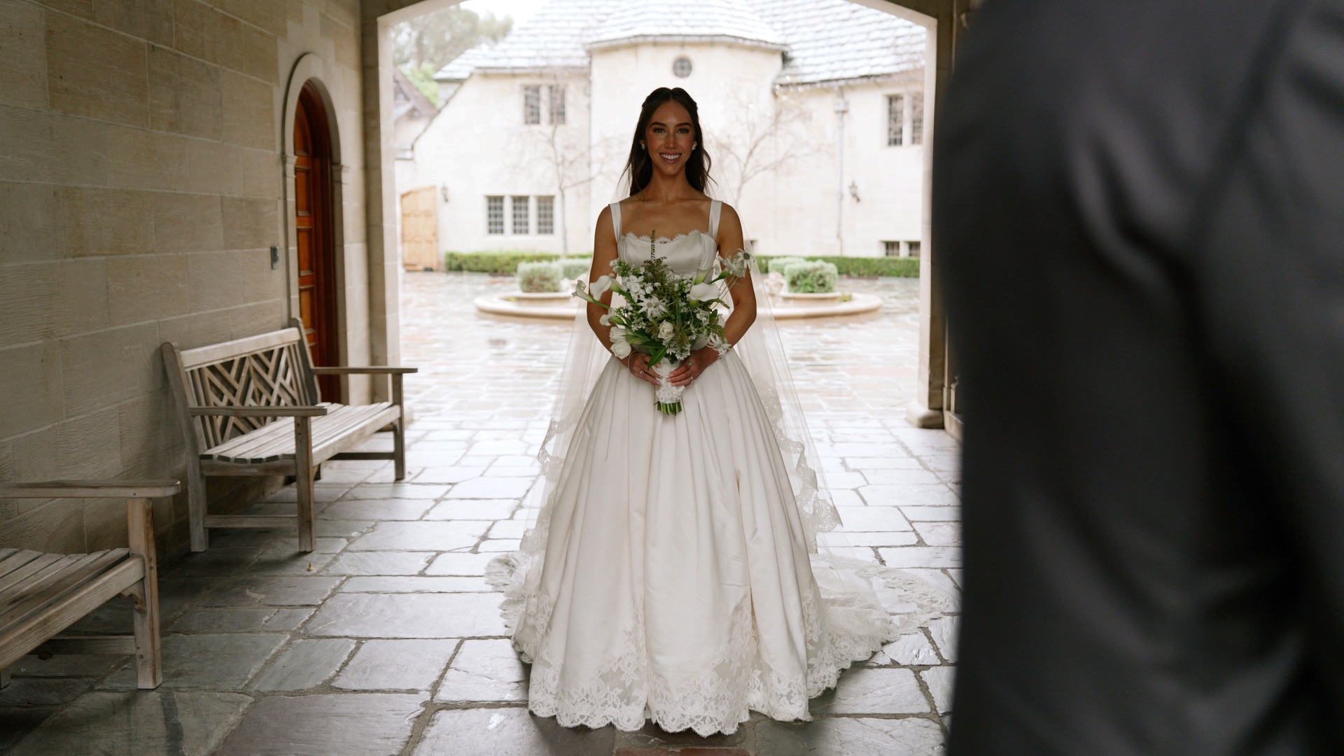 A bride in a satin ballgown and lace-edged cathedral veil before her first look, a rain-soaked stone courtyard behind her