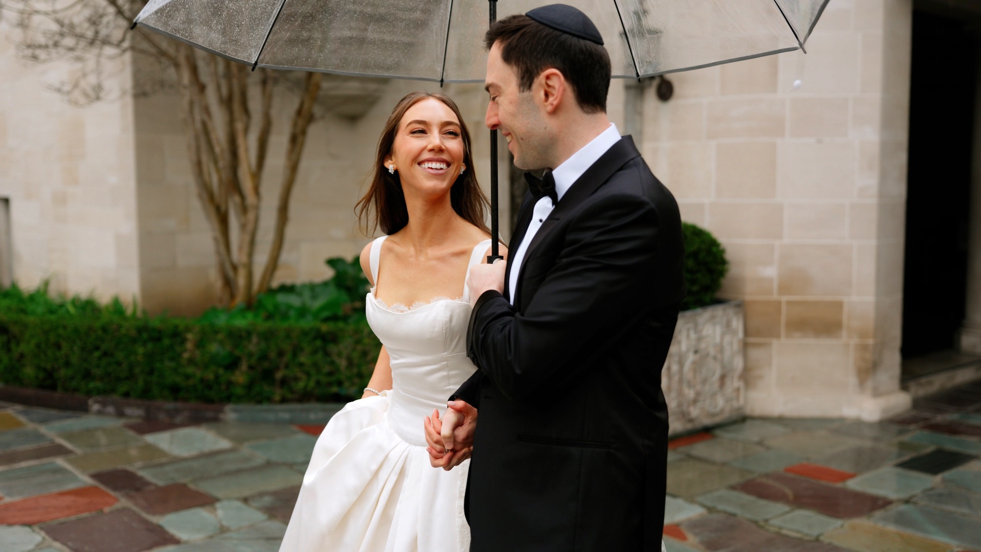 A bride and groom laughing under a clear umbrella in the rain on their Beverly Hills wedding day