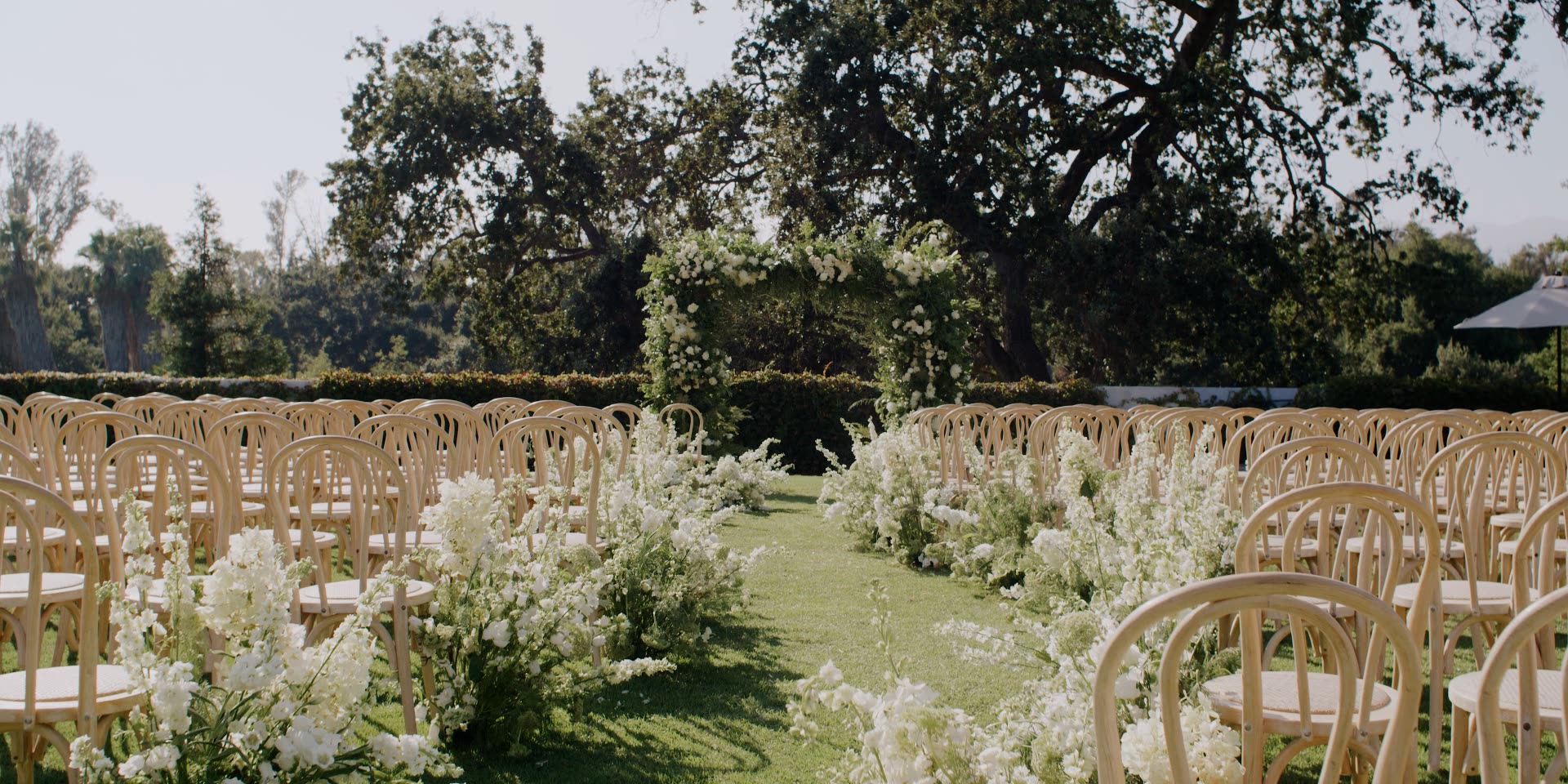 Ojai Valley Inn wedding ceremony on the lawn beneath a coast live oak, cane chairs and a white floral arch