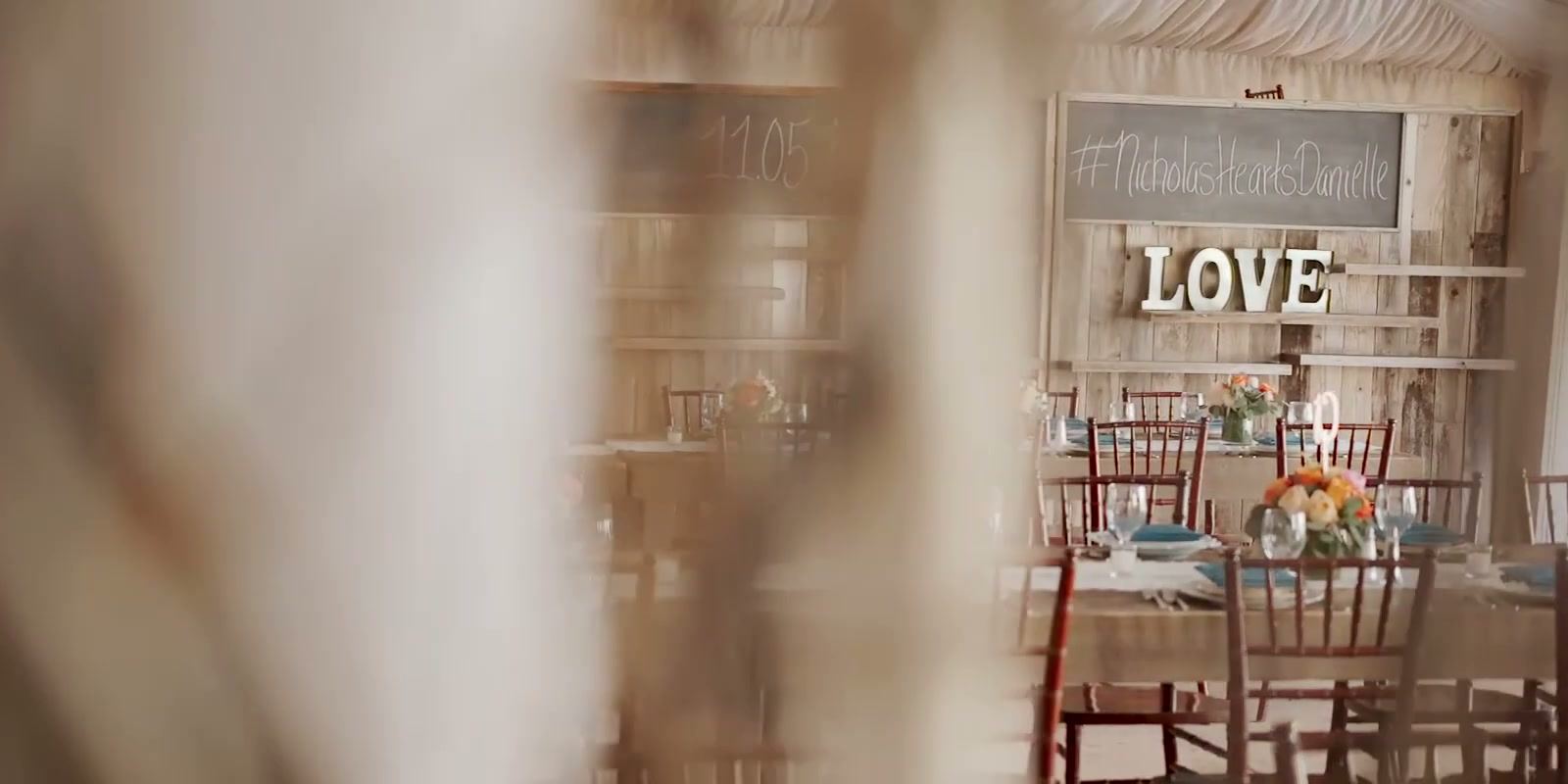 White LOVE letters on a wood wall below a chalkboard with the couple&rsquo;s wedding hashtag, dinner tables set in front.
