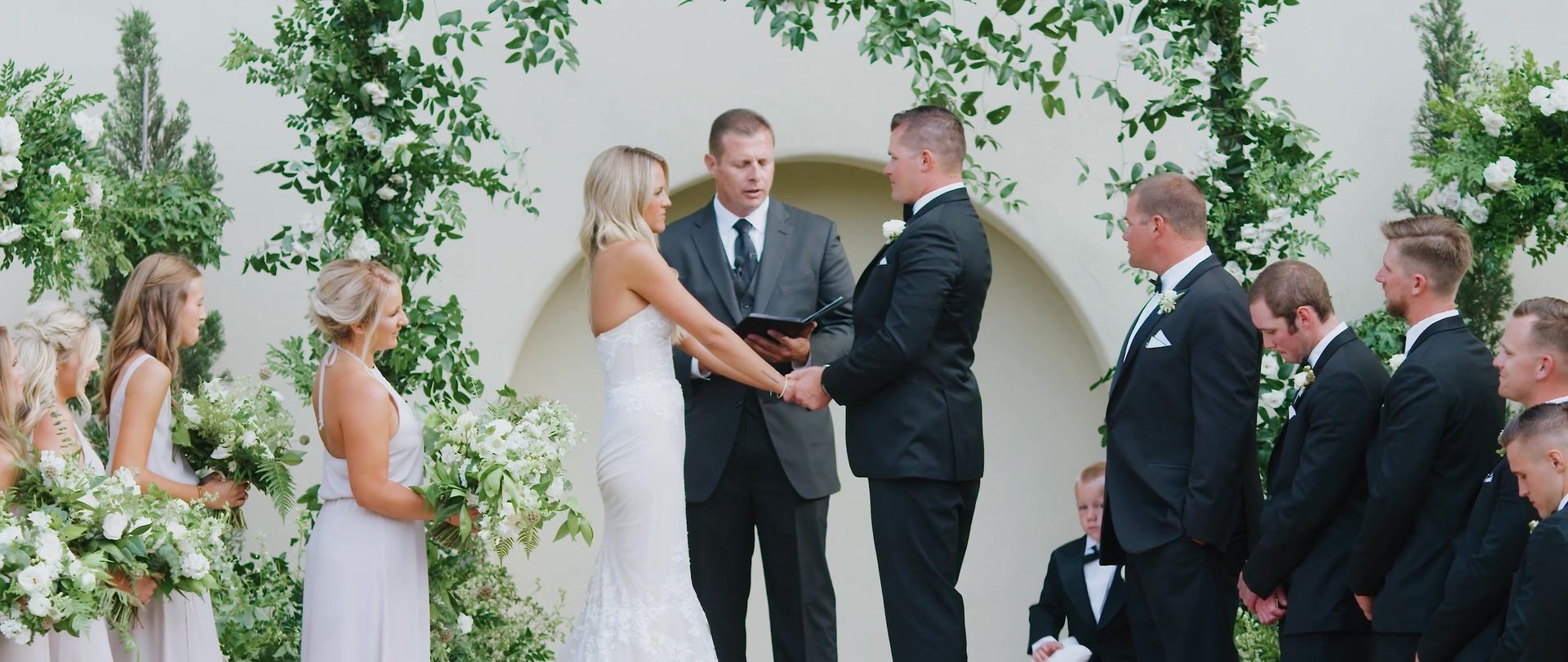 Nick and Paige before an arched recess in the white wall, greenery arches to either side, the wedding party on both sides.