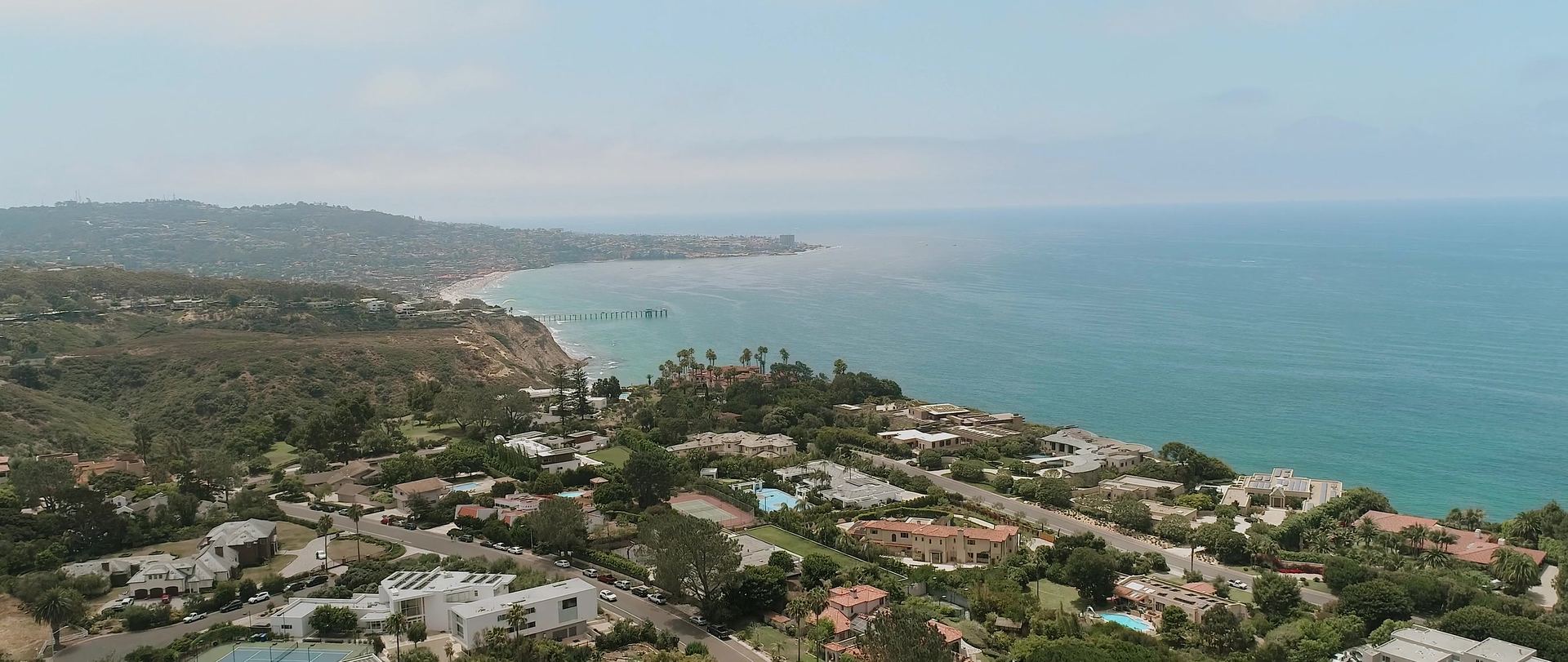 The coastline from above, houses along the bluffs, a pier reaching into the water and a town on the headland beyond.