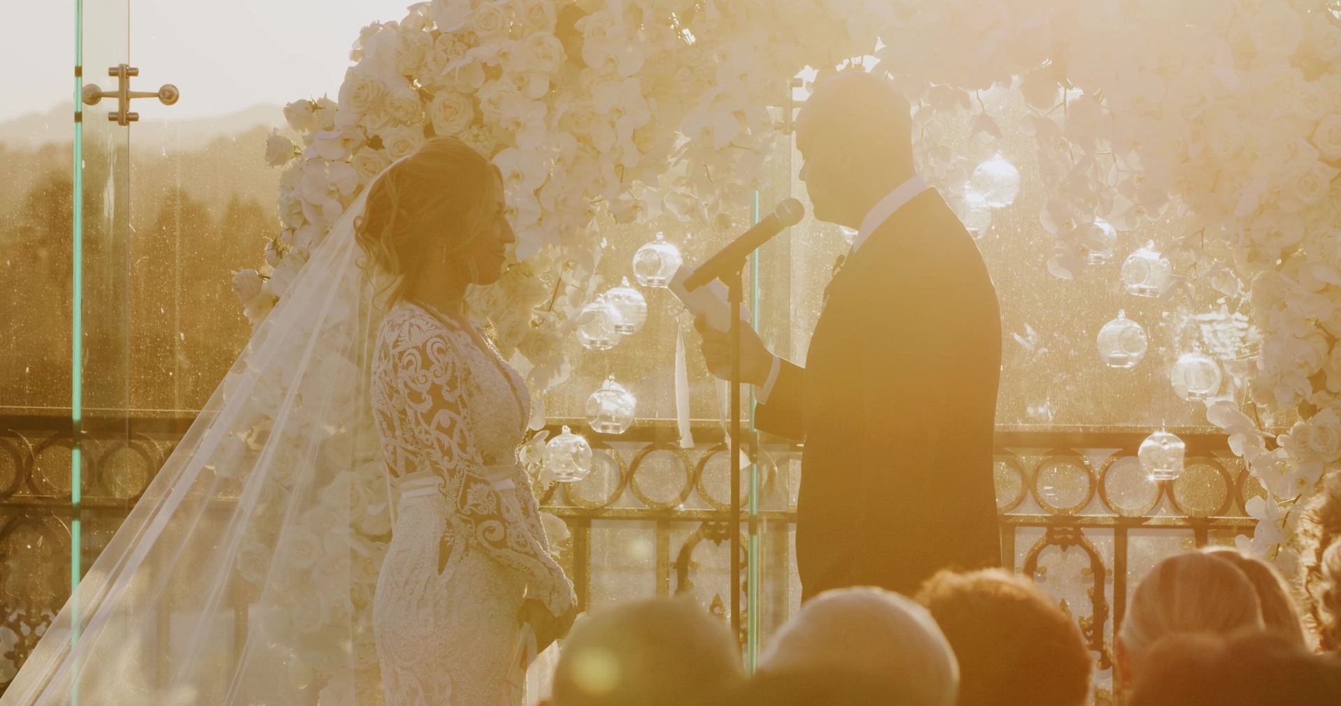 The ceremony backlit through the ring of white blooms, the bride's veil and lace sleeves lit from behind, glass orbs hanging in the arch.