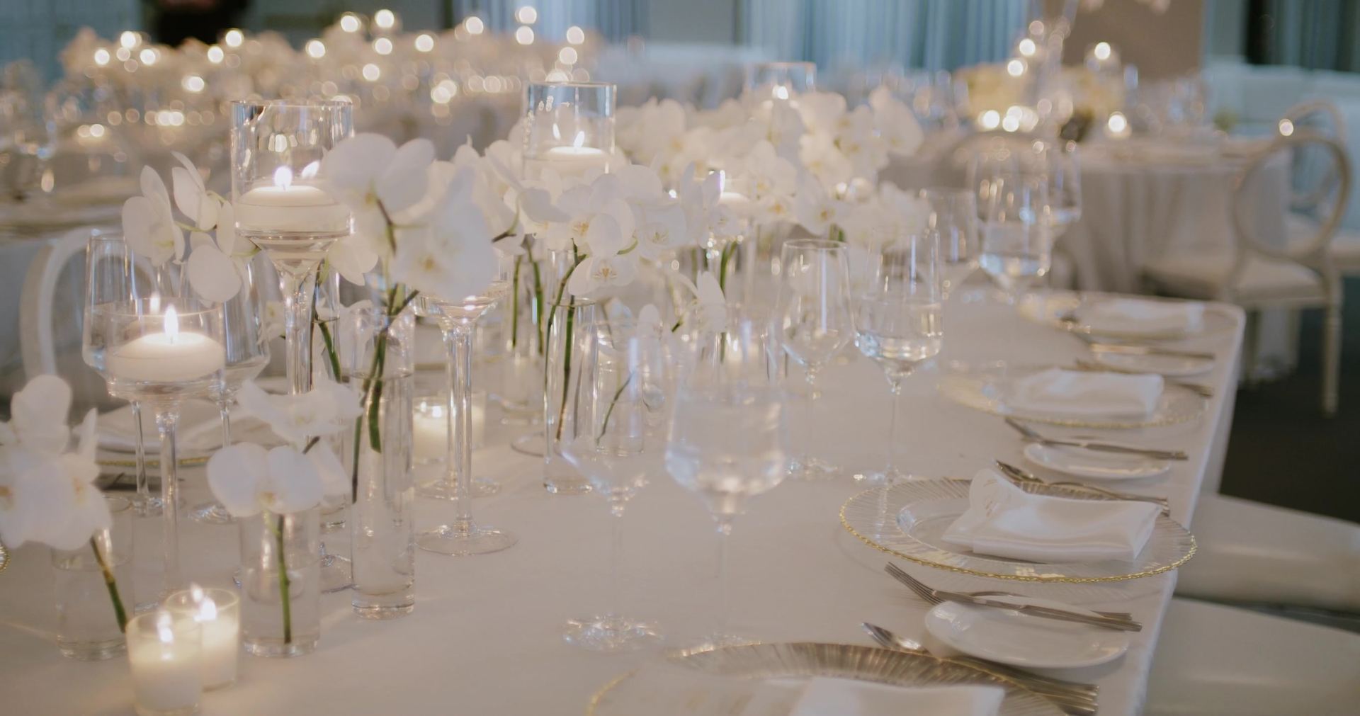 The reception table set all in white, single orchid stems in glass, floating candles and gold-rimmed chargers.