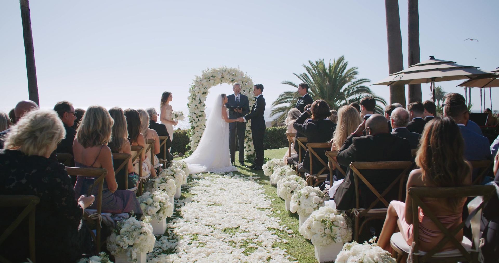 The ceremony on the lawn, a petal-strewn aisle running to a white floral arch, the couple and the officiant at the altar, guests seated in wooden chairs, palms and the ocean beyond.