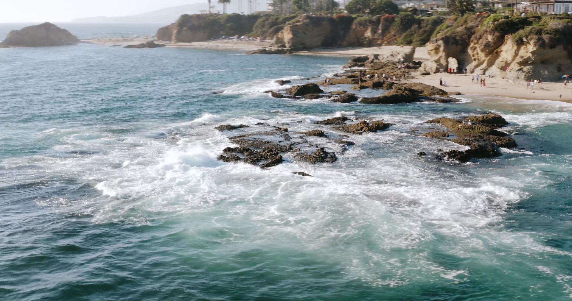 The rocky point and surf from above, a beach and houses along the bluff, people on the sand.