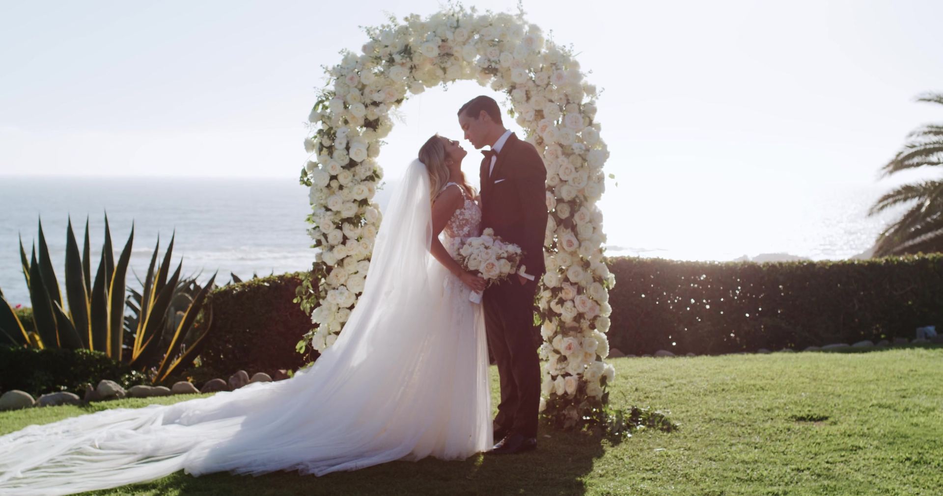 Nic and Sophie beneath an arch of white blooms on the lawn, an agave to the left, her train spread across the grass and the ocean behind.