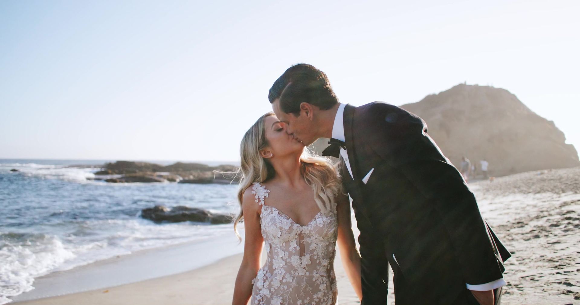 Nic kissing Sophie at the water's edge, a headland and a few figures on the sand behind them.
