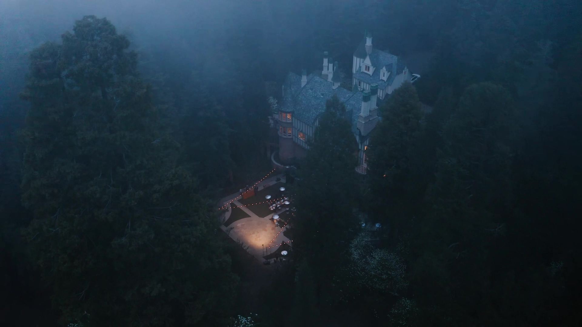 The castle in mist among the pines at night, a few windows glowing and string lights over the terrace below.
