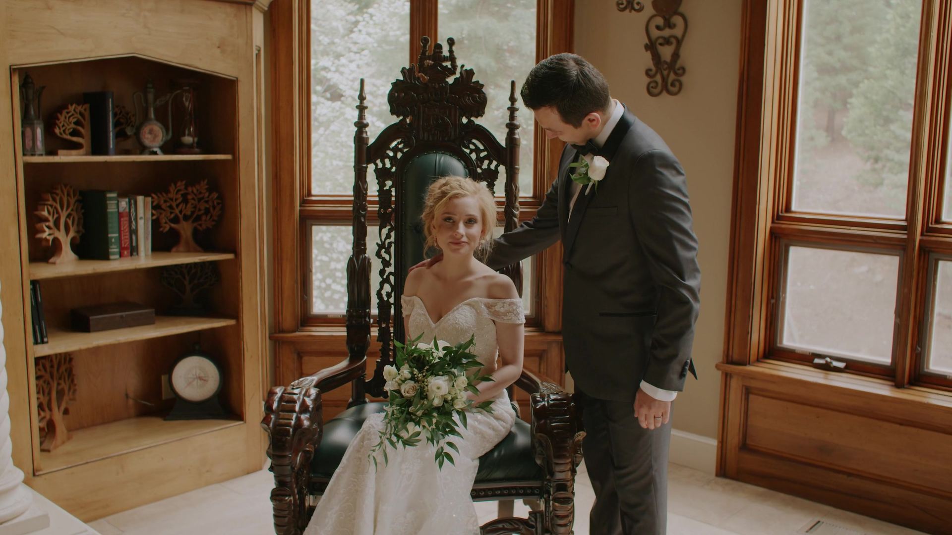 Julya seated in a carved wooden throne chair with her bouquet, Peter standing beside her with a hand on her shoulder, a bookcase and tall wood-framed windows behind.
