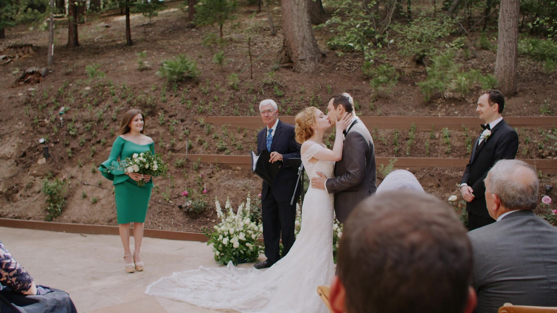 Peter and Julya kissing at the end of the ceremony, the officiant and attendants beside them and the pine hillside rising behind.