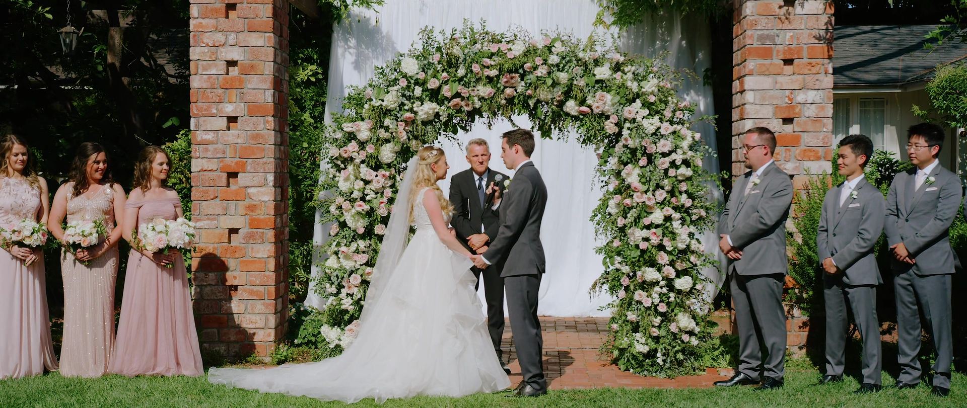 Ryan and Mandi at the altar beneath an arch of white and blush blooms between the brick pillars.